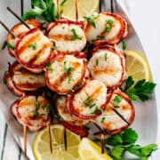 grilled bacon wrapped scallops on skewers on a brushed metal platter.