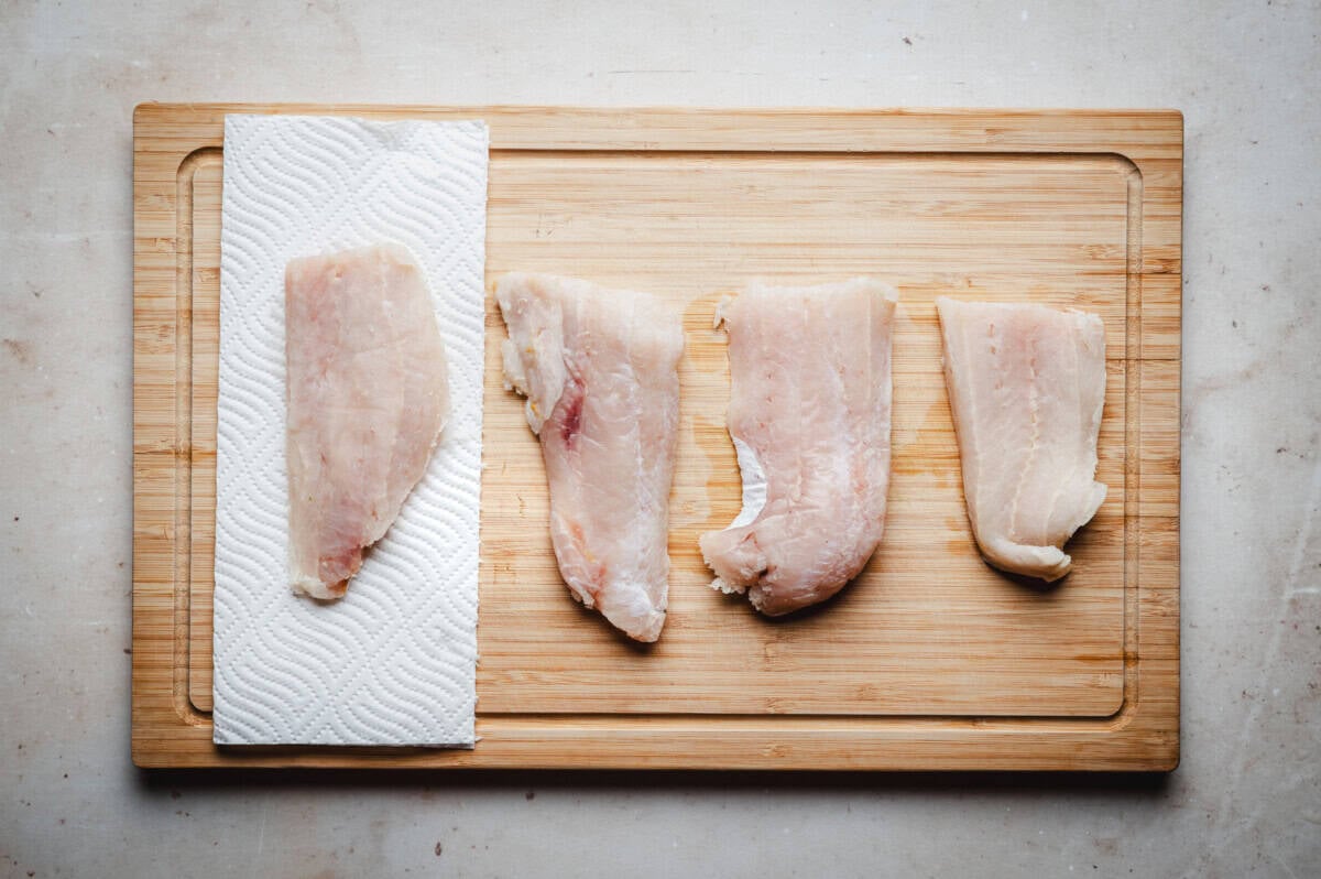 Four raw fish fillets, perfect for cod piccata, are arranged in a row on a wooden cutting board, with one fillet resting on a paper towel to the left. The background features a light-colored surface.