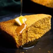 A slice of Sweet Potato Cornbread on a black plate with a pat of butter and drizzle of maple syrup.