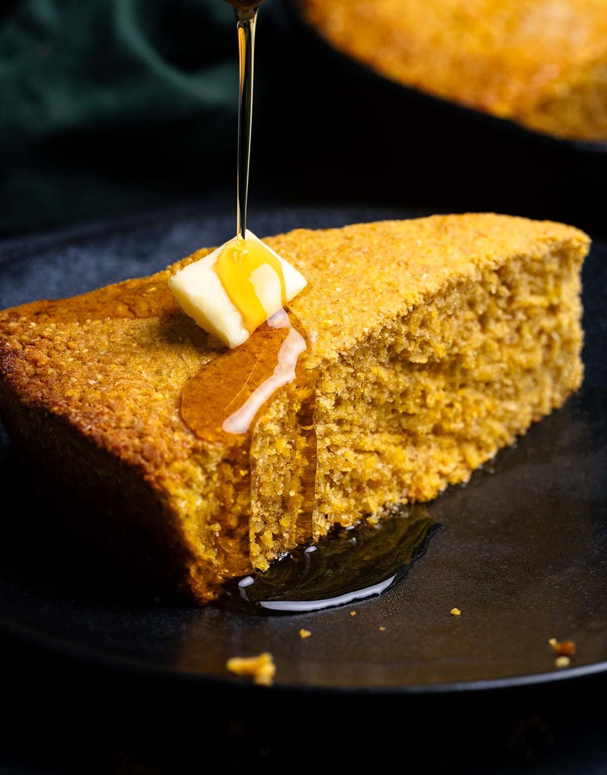 A slice of Sweet Potato Cornbread on a black plate with a pat of butter and drizzle of maple syrup.