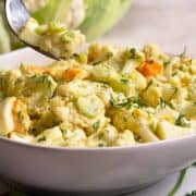 One bite of Cauliflower Potato Salad on a fork from a white serving bowl photographed at an angle.