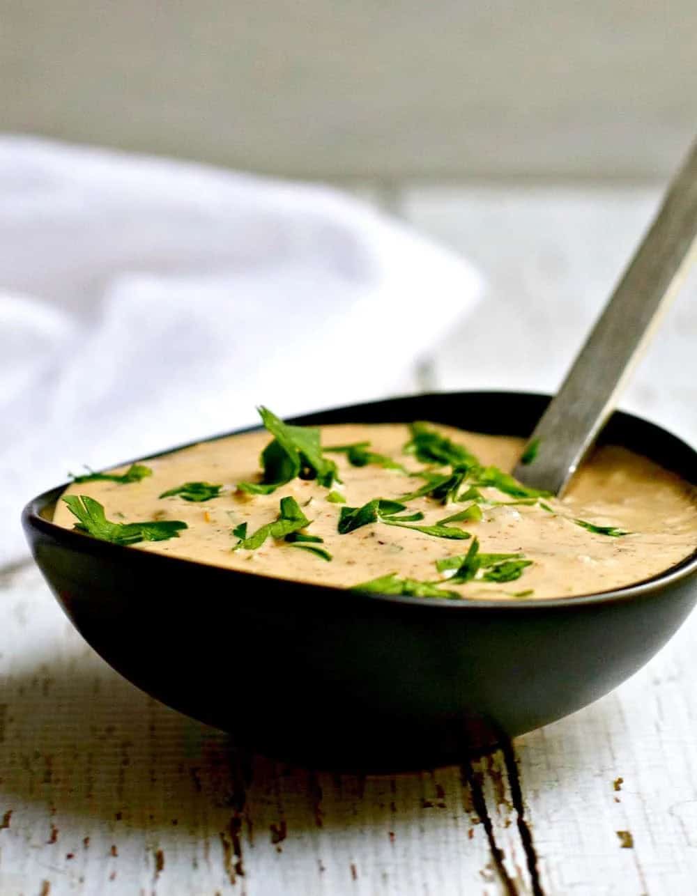 A black bowl filled with creamy remoulade sauce, garnished with chopped fresh herbs, with a spoon resting inside. The bowl is placed on a rustic white wooden surface.