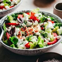 A bowl filled with a colorful Greek Chicken salad, including greens, sliced red bell peppers, cucumbers, olives, capers, and crumbled feta cheese. Tender chicken slices are mixed in, and a creamy dressing is drizzled on top. A fork rests on the side of the bowl.