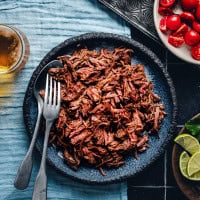 A dark blue plate filled with shredded beef, flanked by a fork and a knife on the left side. Beside the plate are a glass of beer, a bowl of cherry tomatoes, a small dish of lime wedges, and a garnish of fresh herbs on a blue textured surface.