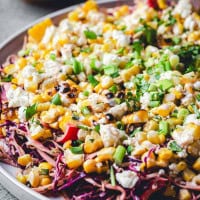 A colorful Mexican Street Corn Salad on a plate, featuring charred corn, crumbled feta cheese, chopped green onions, and red cabbage. The salad is garnished with fresh herbs, and drizzled with a dark balsamic glaze or dressing. The vibrant mix showcases a variety of textures and colors.