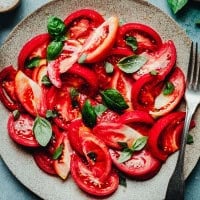 A plate of fresh tomato salad topped with basil leaves and sprinkled with salt. The salad is arranged on a rustic stone plate with a fork placed on the side. The vibrant red of the tomatoes contrasts with the green of the basil, creating a visually appealing dish.