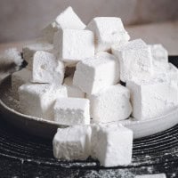 A plate piled with powdered white marshmallows in square shapes, placed on a gray surface. The marshmallows appear soft and fluffy, with some powdered sugar scattered around them.