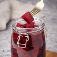 A fork holding up a cube of pickled beets above an open glass jar filled with more beet cubes. The jar has a metal clasp and sits on a gray surface, with a white cloth blurred in the background.