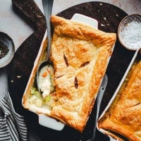 A rectangular baking dish with a golden-brown puff pastry crust, partially removed to reveal a creamy vegetable and chicken filling inside. A spoon rests inside the dish. Surrounding items include a bowl of coarse salt, a small dish with herbs, and a striped cloth napkin.