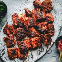 A rustic metal tray holds several skewers of grilled pork, charred and juicy. Visible grill marks enhance the rich, reddish-brown color of the cooked meat. To the side is a spoonful of bright red sauce, and a dark beverage in a glass. The background is a textured grey.