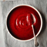 A bowl of smooth, red BBQ Sauce sits on a striped black-and-white cloth. A silver spoon rests on the edge of the bowl, its handle pointing outward. The vibrant color of the soup contrasts with the neutral tones of the cloth and table.