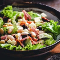 A bowl of fresh salad with large green lettuce leaves, white beans, chunks of cheese, pieces of ham or bacon, and sun-dried tomatoes, served in a rustic dark bowl. The ingredients are evenly mixed, creating a colorful and appetizing dish.