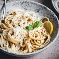 A bowl of creamy spaghetti topped with grated cheese, black pepper, and garnished with a fresh basil leaf and a lemon slice. The dish is served in a speckled, rustic bowl with a fork resting inside.