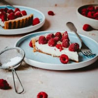 A slice of raspberry tart with a creamy filling on a blue plate, garnished with fresh raspberries and powdered sugar. A metal fork rests on the plate. Additional tart slices, raspberries, a strainer with powdered sugar, and another plate are visible in the background.