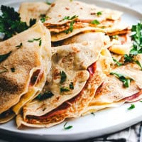 A close-up of a plate stacked with savory crepes, folded and filled with what appears to be ham and cheese. The crepes are garnished with fresh parsley leaves, adding a touch of color to the dish.