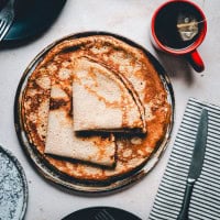 A plate of buckwheat crepes folded in quarters, accompanied by a red mug of tea with a tea bag string hanging out, a knife, and a striped napkin on a light textured table surface.