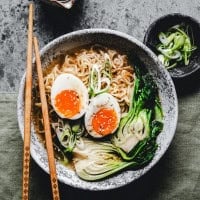 A bowl of ramen with sliced boiled eggs, bok choy, and green onions. Chopsticks rest on the bowl's edge. A small dish with more green onions is beside the bowl on a textured gray surface.