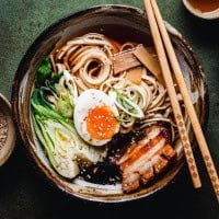A bowl of ramen with noodles, bok choy, a soft-boiled egg, pork belly, bamboo shoots, and green onions, garnished with sesame seeds. Chopsticks rest on the bowl, and small dishes are in the background.