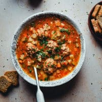 A bowl of hearty albondigas soup with vegetables, garnished with fresh herbs, sits on a light-colored surface. A spoon rests inside the bowl, and slices of rustic bread are placed nearby.