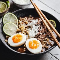 A bowl of birria ramen topped with two halves of soft-boiled eggs, chopped onions, shredded meat, and lime slices. Wooden chopsticks rest on the bowl, placed on a textured surface.