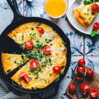 A skillet with a sliced frittata topped with cherry tomatoes and greens. Nearby, a slice on a plate, cherry tomatoes on a cutting board, a knife, and a glass of orange juice sit on a patterned surface.