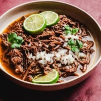 A bowl of savory slow cooker birria stew topped with diced onions, fresh cilantro, and lime slices, set against a rich red broth. The dish is served in a brown ceramic bowl on a textured maroon surface.
