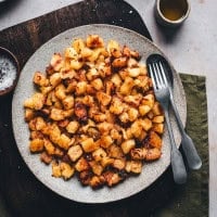 A plate of golden, crispy roasted potatoes on a wooden board, accompanied by silver cutlery. A small bowl of coarse salt and a ceramic cup with dipping sauce are nearby, all placed on a white tabletop.