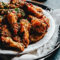 A close-up of a plate of spicy, Cajun seasoned chicken wings garnished with fresh herbs. The wings are crispy and golden brown, sitting on a rustic white plate with a textured cloth beneath.