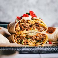 A close-up of a chorizo breakfast burrito cut in half, filled with ground meat, onions, and melted cheese. Topped with pico de gallo, featuring diced tomatoes and onions. The burrito is resting on a piece of parchment paper.