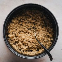 A pot filled with cooked quinoa, with a spoon resting inside. The quinoa is fluffy and light brown in color, and the pot is placed on a light-colored countertop.
