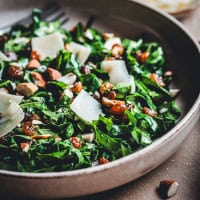 A close-up of a fresh kale salad in a brown bowl. The salad is topped with shaved Parmesan, almonds, and raisins. A fork is partially visible in the background.