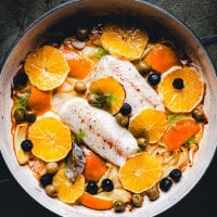A skillet with cooked halibut fish fillets surrounded by sliced oranges, black olives, green olives, herbs, and fennel. The dish is colorful and garnished with fresh green herbs.