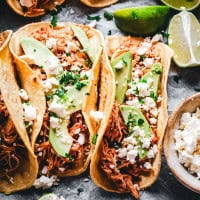Three soft chicken tinga tacos filled with shredded meat, sliced avocado, crumbled cheese, and chopped cilantro. Lime wedges are on the side, with a bowl of extra cheese nearby, all on a rustic surface.
