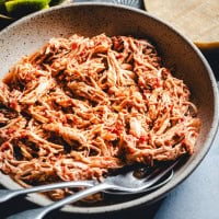 A bowl of shredded chicken tinga, featuring a reddish hue. Two metal forks rest in the bowl, suggesting the dish is ready to be served. There are lime wedges visible in the background.