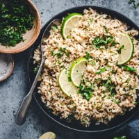 A bowl of seasoned brown rice garnished with fresh cilantro and lime slices. A spoon rests in the bowl, and there is a small bowl of chopped cilantro nearby on a gray surface.