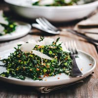 A plate of kale salad topped with shaved Parmesan cheese, set on a wooden table. The salad has a mix of greens and is garnished with breadcrumbs. There are utensils on the plate and a napkin underneath. Additional plates are blurred in the background.