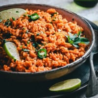 A bowl of vibrant Mexican brown rice garnished with slices of lime, jalapeño pepper, and fresh cilantro. The rice appears fluffy and seasoned, creating a colorful and appetizing presentation.