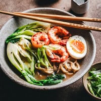 A bowl of shrimp ramen topped with shrimp, bok choy, green onions, and half a soft-boiled egg. The broth is rich, with sesame seeds sprinkled on top. Chopsticks rest on the bowl's edge, and a small cup is in the background.
