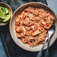 A bowl of shredded chicken seasoned with spices and herbs. The dish is placed on a textured surface, alongside a small bowl containing lime wedges. Two utensils are partially visible in the dish.