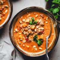 A bowl of creamy West African peanut stew garnished with peanuts, cilantro, and black pepper. The stew has chunks of vegetables and a rich, reddish-brown broth. A spoon rests in the bowl, and a sprig of fresh cilantro lies beside it on the table.