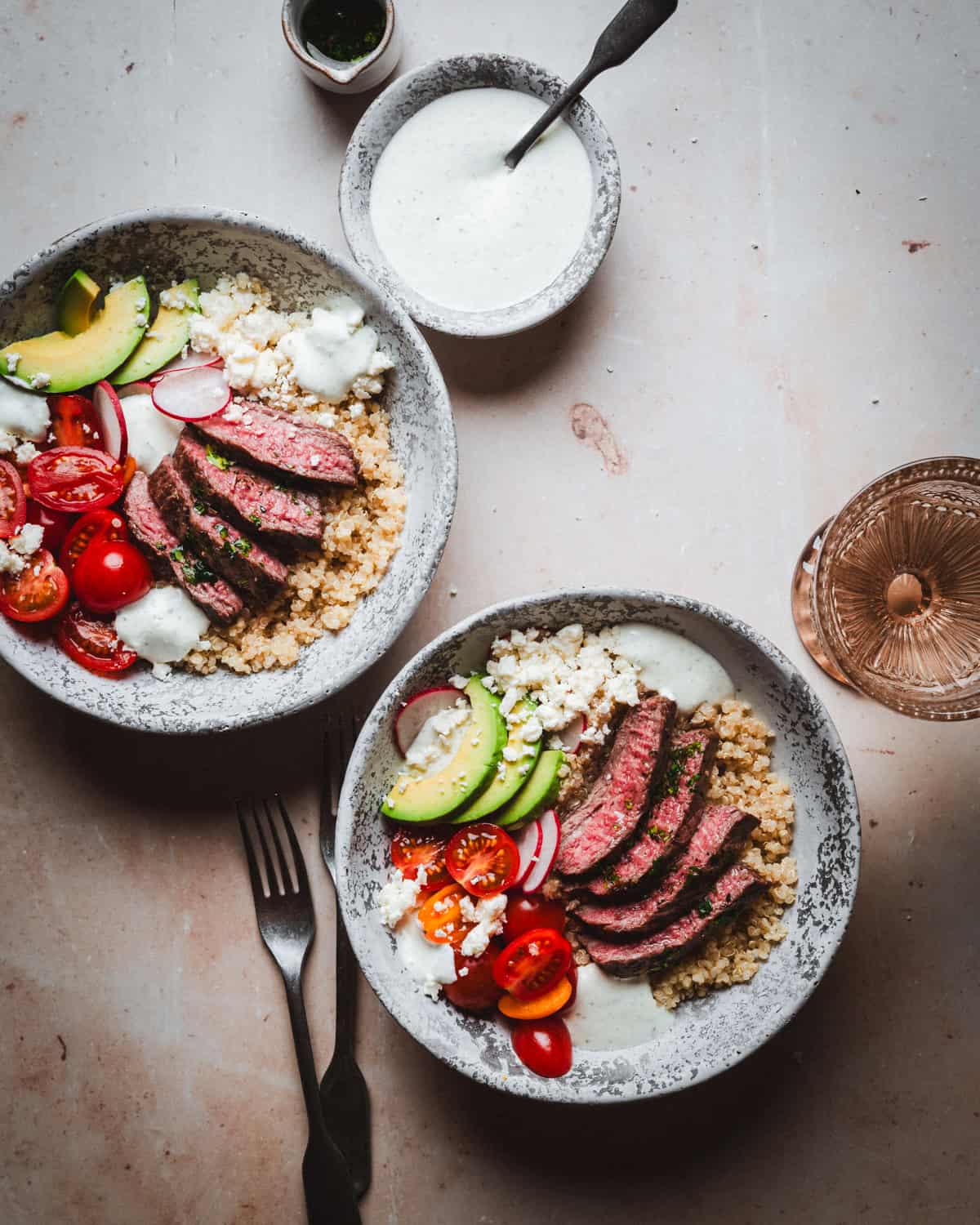 Two bowls of quinoa topped with sliced steak, avocado, cherry tomatoes, and radishes, garnished with creamy dressing and crumbled cheese. A small cup of dressing is nearby on a light marble surface.