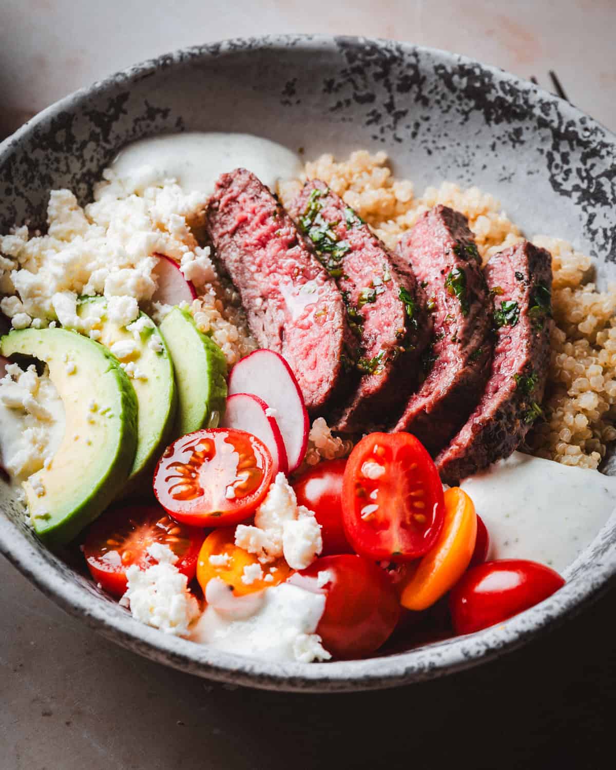 A bowl filled with slices of steak, quinoa, avocado, cherry tomatoes, radish slices, and crumbled cheese, all drizzled with a creamy dressing. The ingredients are arranged in a visually appealing, colorful presentation.