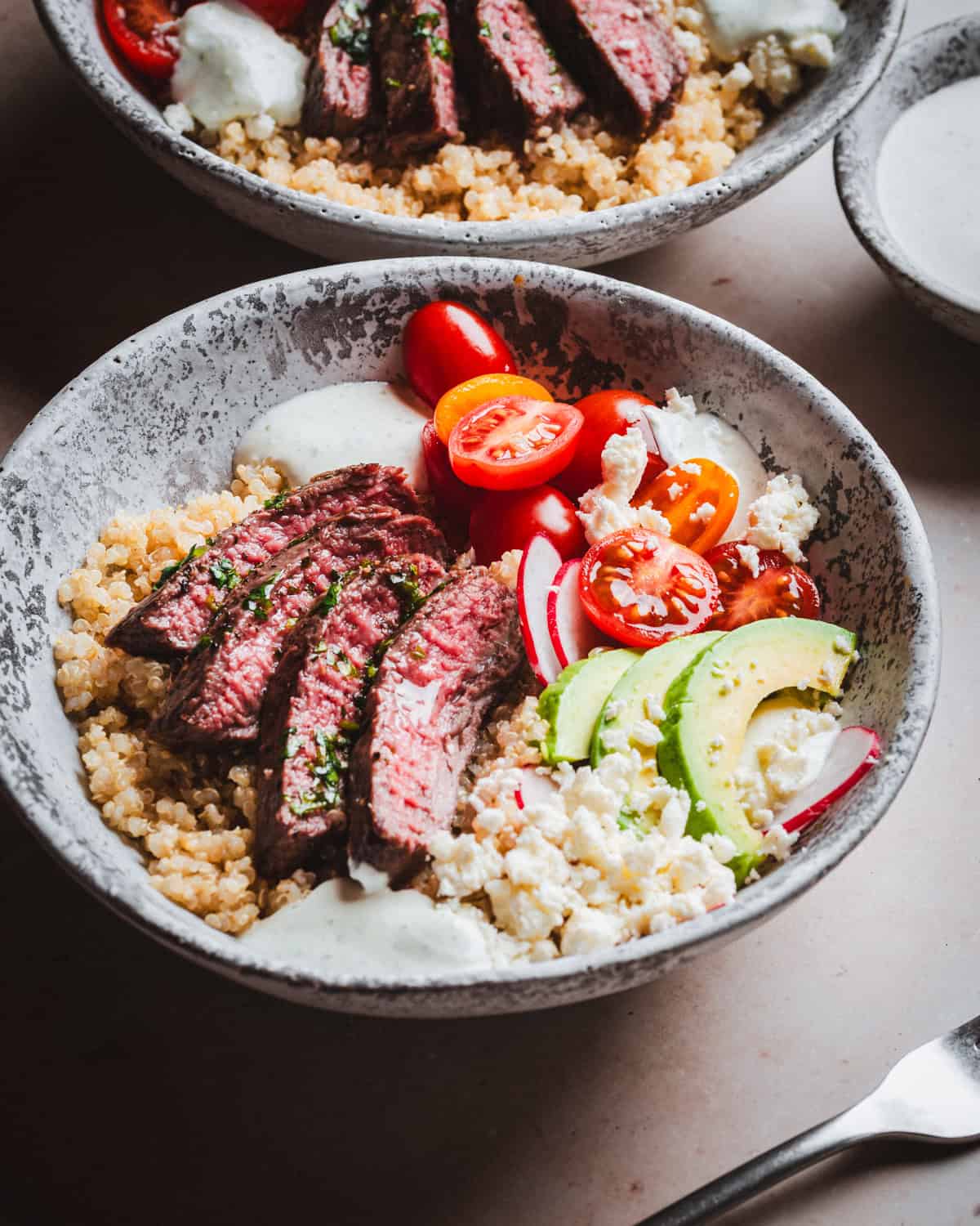 A bowl filled with sliced grilled steak, quinoa, cherry tomatoes, avocado slices, radish, crumbled cheese, and a drizzle of creamy dressing. The ingredients are artfully arranged, creating a colorful and appetizing dish.