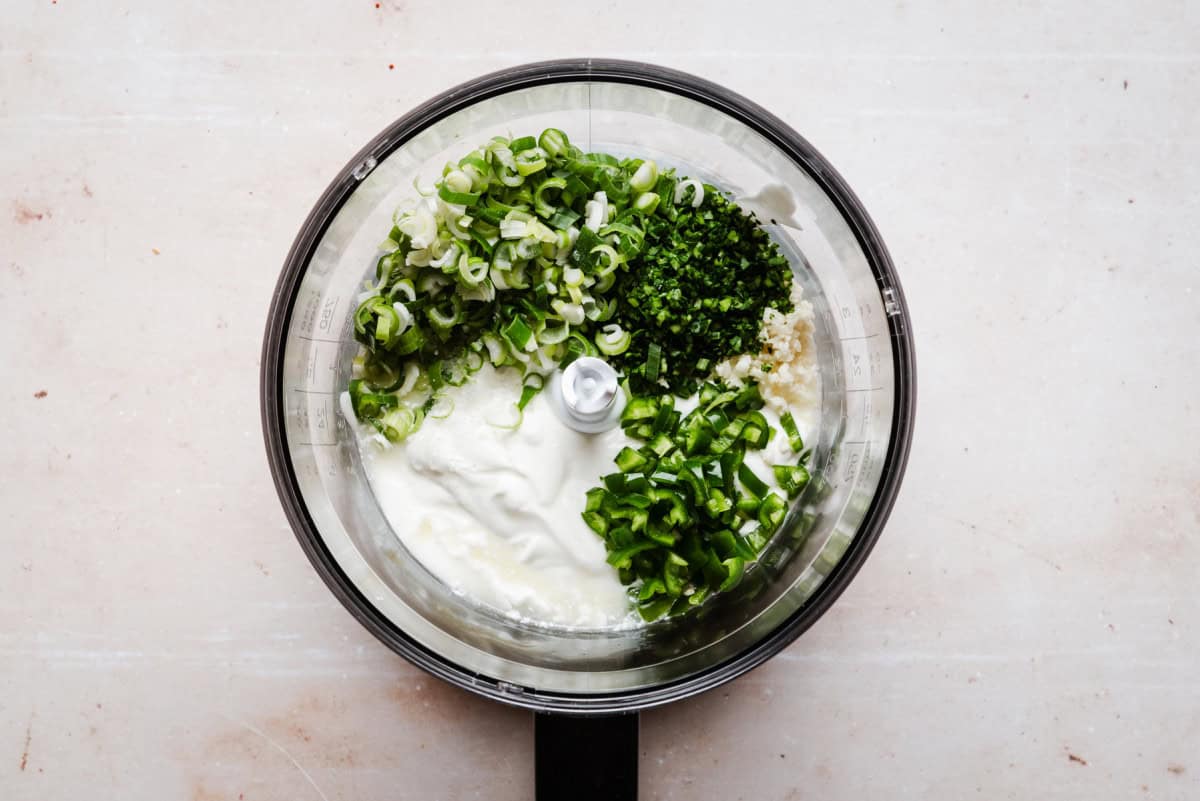 Overhead view of a food processor containing chopped green onions, cilantro, garlic, sour cream, and green chilies. The ingredients are ready to be blended on a light-colored countertop.