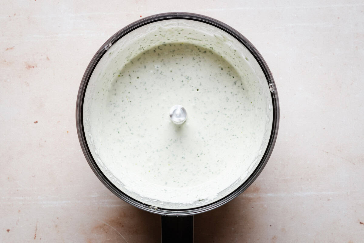 A top view of a food processor bowl containing a creamy, white dressing with green onions and cilantro blended in, placed on a light-colored countertop.