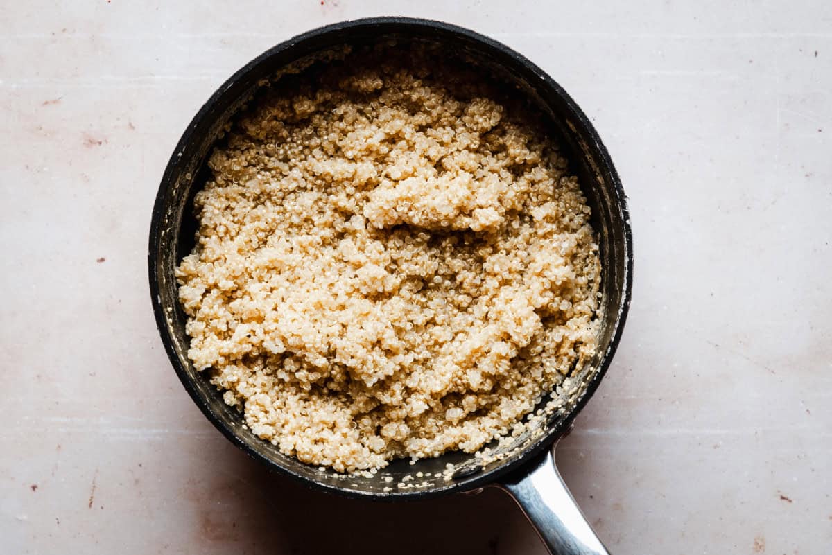 A black saucepan filled with cooked quinoa on a light beige countertop. The quinoa has a fluffy texture and a light golden color. The saucepan has a metallic handle, positioned to the right.