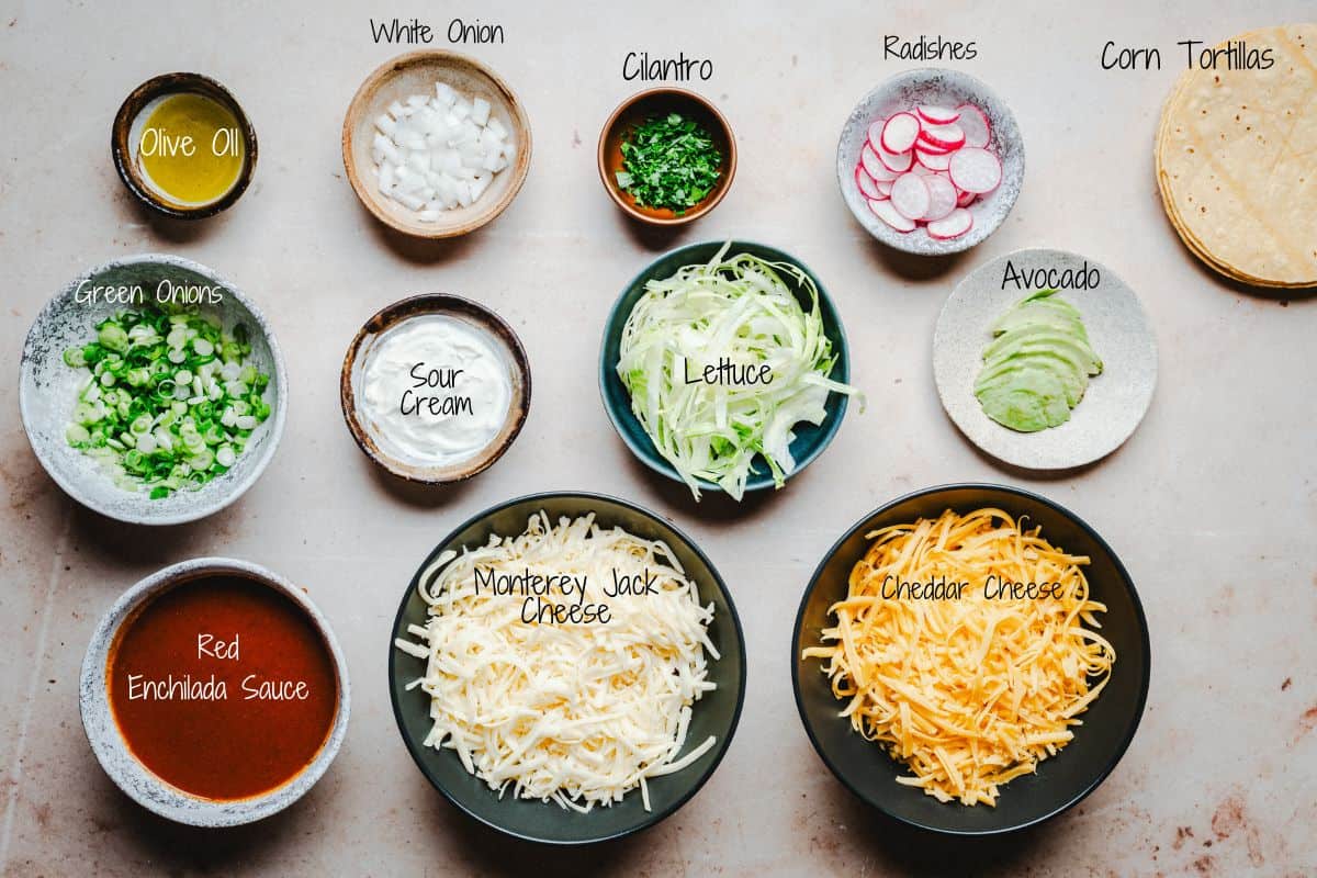 Cheese enchilada ingredients are arranged on a table, including chopped white onion, cilantro, radishes, corn tortillas, olive oil, green onions, sour cream, lettuce, avocado, red enchilada sauce, Monterey Jack cheese, and cheddar cheese.