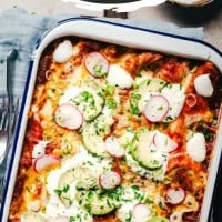 Overhead view of a baked dish of cheese enchiladas topped with sliced radishes, sour cream, and chopped green onions, served in a rectangular baking dish. A folded gray napkin and cutlery lie beside it. "Cheese Enchiladas" text is visible at the top.