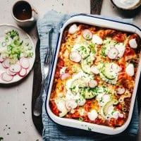 A baking dish filled with cheese enchiladas topped with cheese, avocado slices, radish, and green onions. Beside it are a fork, a knife, a gray napkin, a bowl of sauce, and a small plate with sliced radishes and green onions.