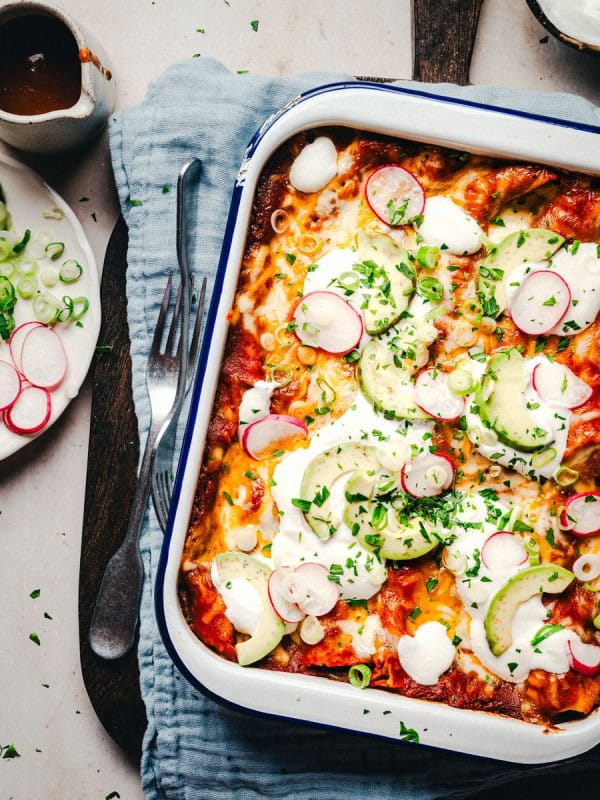 A baking dish filled with cheese enchiladas topped with cheese, avocado slices, radish, and green onions. Beside it are a fork, a knife, a gray napkin, a bowl of sauce, and a small plate with sliced radishes and green onions.